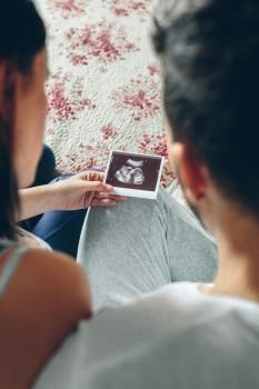 Couple looking at ultrasound of their baby sitting on the bed. Selective background focus on ultrasound. Couple looking at ultrasound of their baby
