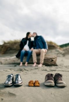 Family shoes in the sand with pregnant woman in the background kissing her partner. Selective focus on shoes in foreground. Family shoes in the sand with couple kissing