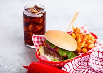 Fresh beef burger with sauce and vegetables and glass of cola soft drink with potato chips fries in red serving basket on stone kitchen background. 