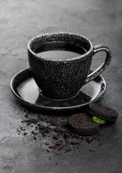 Black coffee cup with saucer and black sandwich cookies on black stone kitchen table background.