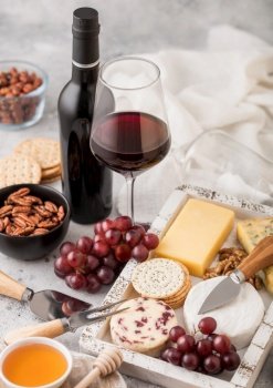 Bottle and glass of red wine with selection of various cheese in wooden box and grapes on light background. Blue Stilton, Red Leicester and Brie Cheese with Cheddar and knife.