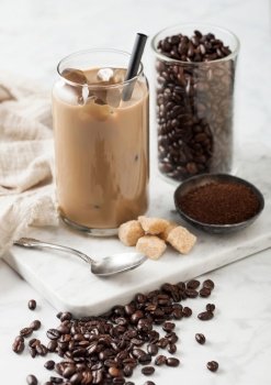 Iced cold caramel coffee with milk and raw beans and spoon with ground coffee and cane sugar on marble board on light.