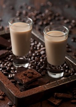 Irish cream baileys liqueur in shot glasses in wooden tray with coffee beans and powder with dark chocolate on dark wood background. Macro