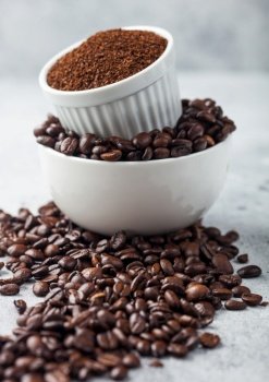 Two bowls on top of each other with fresh raw coffee beans and powder on light table background.