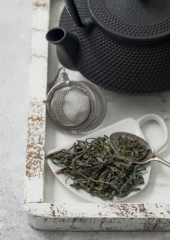 Loose green tea on cup shape ceramic plate and tea ball strainer infuser with iron teapot in white wooden box. Macro