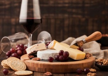 Glass of red wine with selection of various cheese on the board and grapes on wooden table background. Blue Stilton, Red Leicester and Brie Cheese and knife.