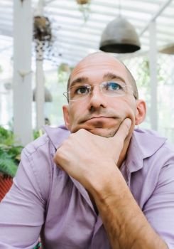 Portrait of an attractive thinking bald man wearing glasses in a pensive mood, holding his chin with the hand and looking up. Soft selective focus.. Attractive Pensive Bald Man Wearing Glasses