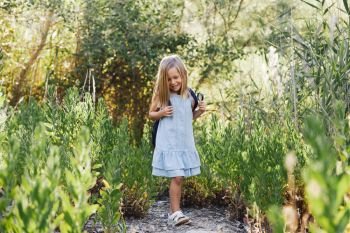 Young blonde girl walking in the field wearing a dress with a magnifying glass in her hand an backpack