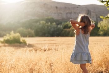 Young blonde girl playing with sunlight in the fields sunset wearing a dress with hill in the background