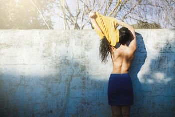 Beautiful brunette against the wall of an empty pool undressing with the sun illuminating her naked back