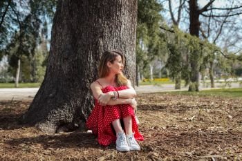 Young serious blond woman siting near tree with red long dress