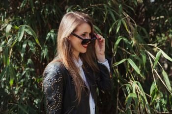 Young smiling blonde woman wearing a leather jacket in the park with sunglasses