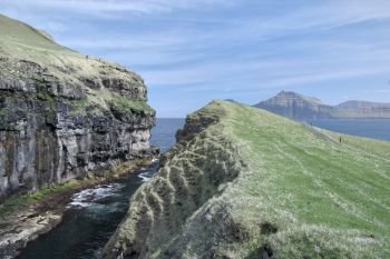 Horizontal scenery image to the sea-filled gorge nearby idyllic village Gjogv, most northern village on the island of Eysturoy in foreground surrounded by high cliffs. Glorious sceneries of the Faroes. Postcard motif.