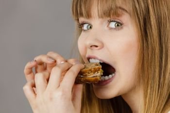 Young woman eating sandwich, taking bite with wide open mouth. Food, calories, dieting concept. Studio shot on grey background, profile view.. Woman eating sandwich, taking bite