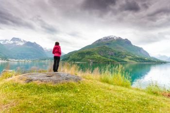 Travel concept. Mature tourist woman on sea shore looking at fjords beautiful landscape, in Olden village, Sogn og Fjordane county Norway.. Woman tourist relaxing on fjord sea shore, Norway