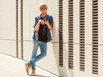 Men fashion, technology, urban style clothing concept. Hipster guy standing on city street wearing jeans outfit and weird sunglasses listening to music and looking at phone. Hipster man listening music through earphones