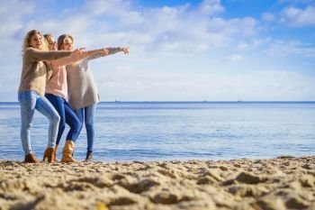 Three fashionable women wearing sweaters during warm autumnal weather pointing at copy space on sunny beach. Fashion models outdoor. Three fashionable models pointing outdoor