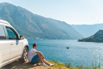 young man sitting near suv car at seaside with beautiful view of sea bay with mountains. road trip. young man sitting near suv car at seaside with beautiful view of sea bay with mountains