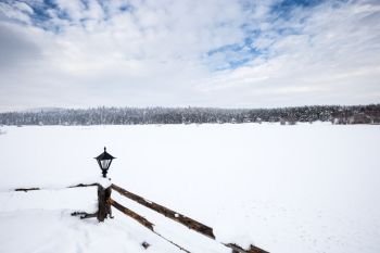 Winter landscape view with pine forest at a cloudy dull day.Fence and a small lamp on the foreground.. Winter landscape view with pine forest