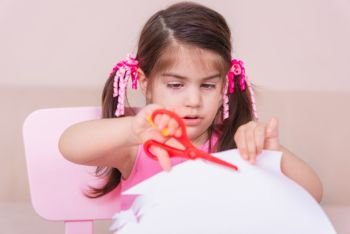 Portrait of Cute girl cuts paper with a red scissors while sitting at pink table. Selective focus and small depth of field.. Portrait of Cute girl cuts paper with a red scissors 