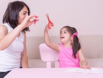 Mother teaches her daughter how to use scissors and cut paper while sitting at pink table. Selective focus and small depth of field.. Mother teaches her daughter how to use scissors