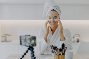 Happy female beauty blogger records video for her blog while applying beauty moisturising patches under eyes wears wrapped towel on head and domestic robe shares opinion about cosmetic product