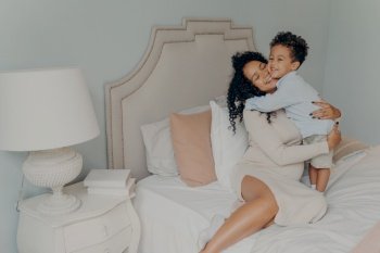 Happy family at home. Smiling pregnant mixed race woman mother in dress with curly hair hugging her beloved sweet little son while sitting on big soft bed. Mom and little child spending time together. Cute little son hugging his pregnant mom, waiting for future brother or sister