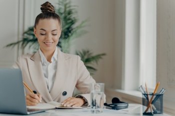Young successful businesswoman in formal wear enjoying office work, writing down information in notepad with positive expression while sitting at workplace with laptop. Job and occupation concept. Young successful businesswoman in formal wear writing down information in notepad