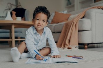 Little cutie curly haired afro baby boy sitting on floor of modern living room interior next to sofa and drawing with colorful felt tip pens in album. Creative hobby for kids and playtime activity. Little afro kid boy sitting on floor and drawing with colorful felt tip pens