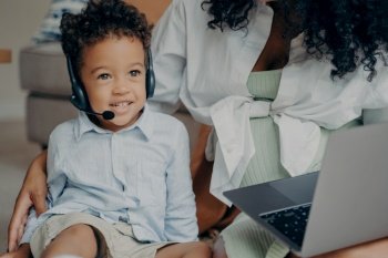Happy afro american child with curly hair looking aside and smiling, wearing headset while having online meeting with dad, using laptop with his mother, boy talking with family through webcam. Happy afro american boy talking with family through webcam on laptop with mothers help