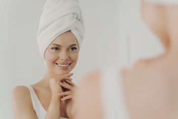 Positive healthy young woman smiling while applying facial cream reflecting in mirror, happy attractive lady putting moisturizing nourishing cream during morning beauty routine in bathroom. Healthy young woman with clean skin smiling while applying facial cream