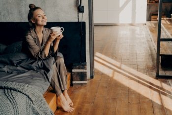 Starting new beautiful day. Cute pretty woman with cup of morning coffee chilling while sitting on bed, relaxing in brown satin pajama in modern apartment. Happy time at home. Cute pretty girl with cup of morning coffee chilling while sitting on bed