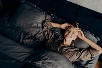 Image of young beautiful sleepy girl in satin comfy pajama waking up stretching in early morning in comfy bed rejoicing in new day, happy and broadly smiling while looking up. Beautiful young girl in satin comfy pajama waking up while lying in bed at home