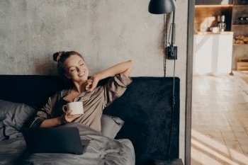Happy beautiful female keeping eyes closed in satin pajama holding cup of coffee and enjoying morning sun, lying in bed with laptop after waking up in morning before starting working remotely at home. Happy beautiful female with closed eyes drinking coffee in bed before remote work at home