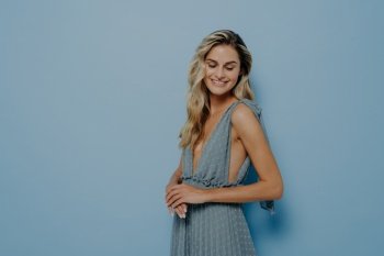 Studio portrait of young smiling beautiful female model with long blonde wavy hair in soft blue evening dress with deep neckline posing sideways on pastel blue background with copy space. Young smiling beautiful female model in blue evening dress posing sideways on pastel blue background