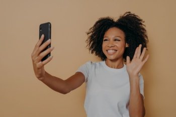 Hello! Beautiful cheerful dark skinned woman smiling at camera on modern smartphone, taking selfie on mobile phone and feeling happy, gesturing hi while making video call, isolated over studio wall. Cheerful african woman gesturing hi while making video call or taking selfie on smartphone