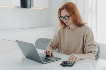 Redhead European female accountant analyzes documents uses calculator makes report uses laptop computer sits at white desktop wears spectacles beige jumper poses against white home interior.. Redhead European female accountant analyzes documents uses calculator makes report