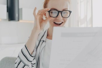 Positive redhead young woman smiles broadly wears transparent glasses focused at papers checks financial report glad to finish work in time works from home. People paperwork and job concept.. Positive redhead young woman smiles broadly wears transparent glasses focused at papers
