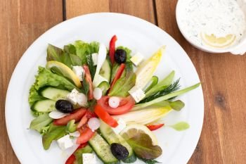 Greek salad with feta, tomatoes, cucumber, peppers and black olives. With bowl of sauce on a wooden table.