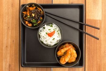 Asian vegetarian dish composed with three black bowls with shrimps, rice noodles, fried vegetables and chinese chopsticks on a square plate. Composition on a old styled wooden table.