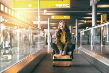 the smiling happy girl is riding on the travolator at the airport with a large yellow suitcase 
