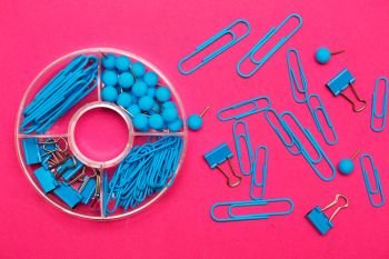 stationery paper clips, binders and buttons in blue on pink background
