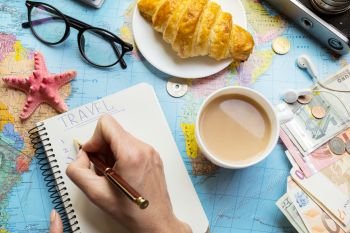 Plan of trip. Women’s hands and notepad for writing ideas, map, retro camera, money, coins, croissant, coffee, pen, sunglasses, smartphone

