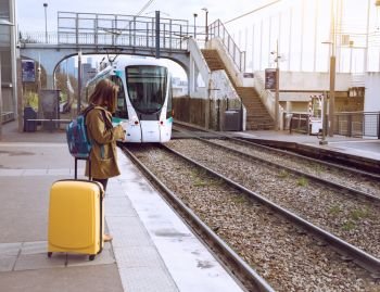 girl tourist with a backpack and a big yellow suitcase stands on the platform and waits for the train
