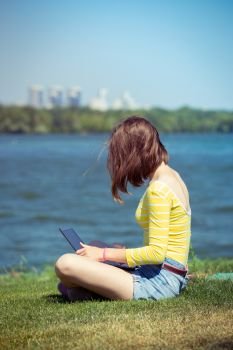 teen girl with a laptop sitting on a  meadow near the river
