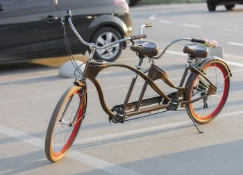 A chocolate-colored tandem bicycle with scarlet wheels is parked in a car park near the bike path in the evening sun.. A tandem bicycle with scarlet wheel rims is parked in a parking lot in the evening sun.