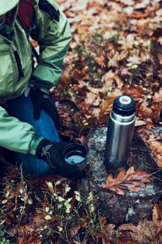 Woman with backpack having break during trip on autumn cold day drinking a hot drink from thermos flask. Active middle age woman wandering around a forest actively spending time. Woman with backpack having break during trip on autumn cold day drinking a hot drink from thermos flask