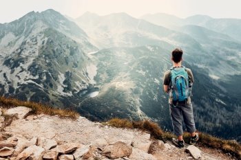 Young man with backpack hiking in a mountains, actively spending summer vacation. Rear view of man standing on the top of a hill admiring mountain landscape panorama. Young man with backpack hiking in a mountains, actively spending summer vacation
