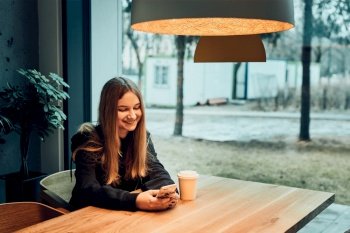 Young woman having a video call, talking remotely, drinking coffee, sitting in a cafe. Girl relaxing in cafe using mobile phone