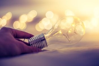 LED light bulb is lying in the bed, hands touching. Symbol for ideas and innovation. Spot lights in the blurry background.
